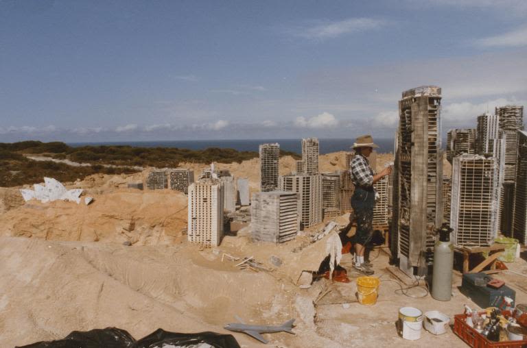 Sydney miniature under construction, woman works on building a skyscraper on the righthandside, Opera House engulfed in sand on the lefthandside